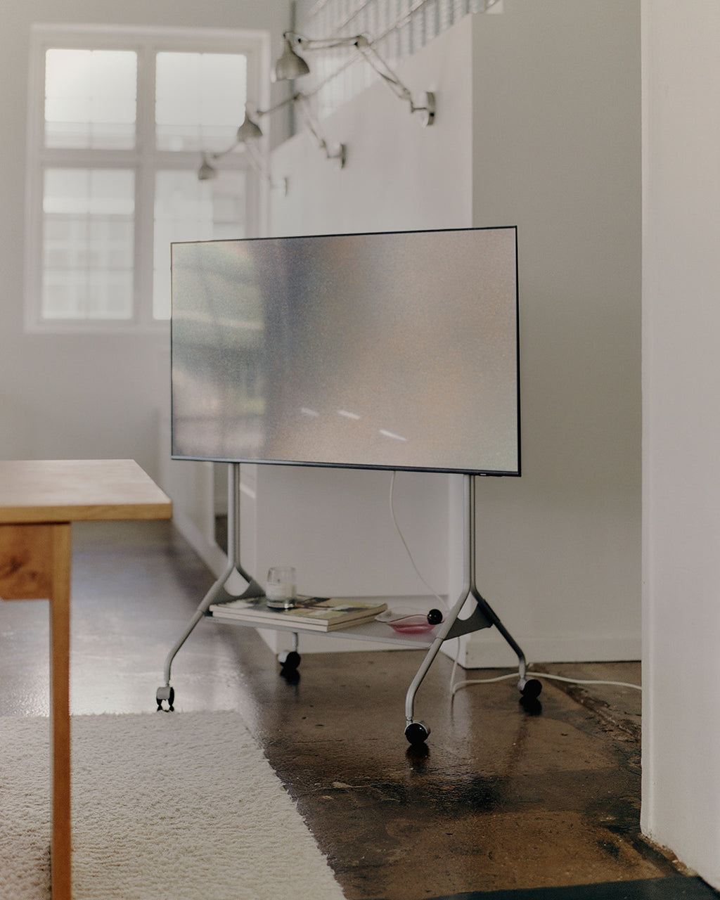 Moon Tall en Cloudy Silver con ruedas y TV montado en sala luminosa, con estante inferior y mesa de madera al lado