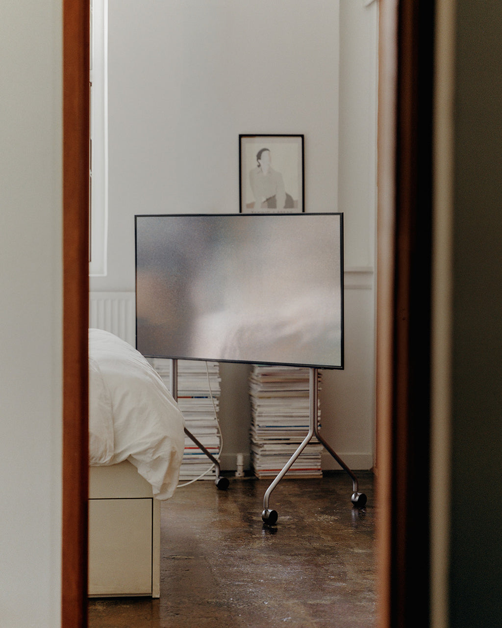 Moon Tall en Cloudy Silver con TV montado y ruedas, visto desde el pasillo en un dormitorio con revistas apiladas